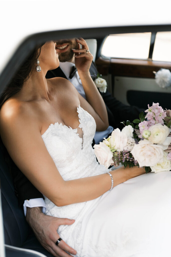 Bride and groom sitting close to each other in back seat of a car, bride's hand on his cheek.