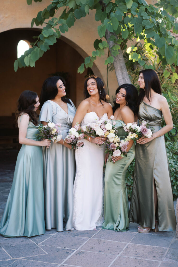 Bride smiling with bridesmaids standing in a line. Bridesmaids wearing shades of green dresses.