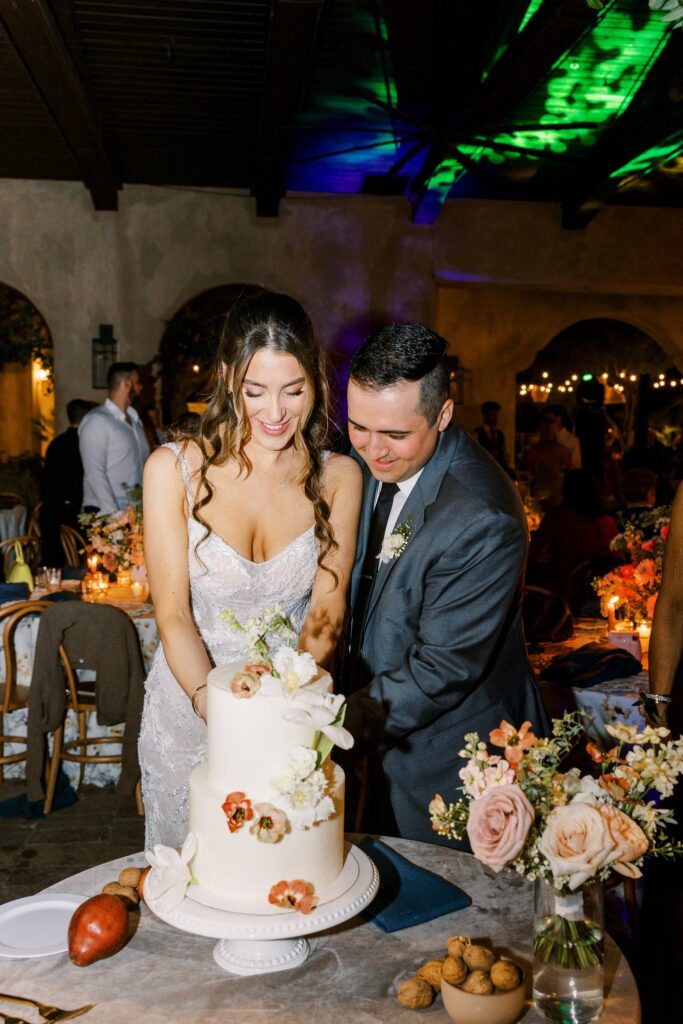 Bride and groom cutting two tiered white cake with flowers added to outside.