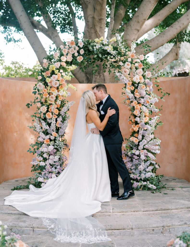 Bride and groom kissing under wedding arch with floral installation of ombre purple, peach, pink, and white flowers and greenery.