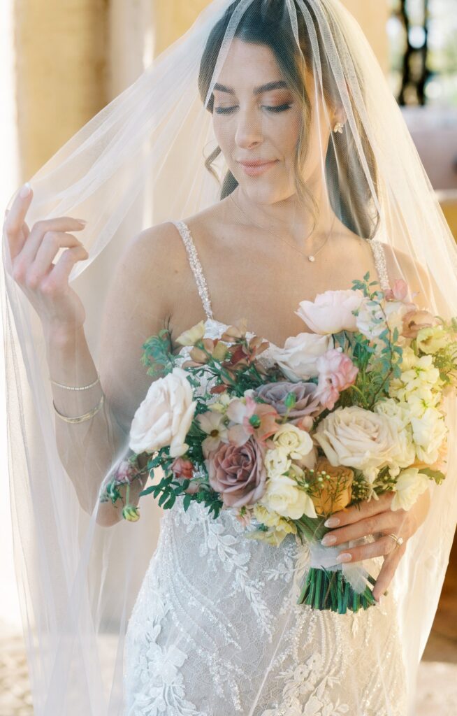 Bride looking down and to side with eyes closed holding bouquet and veil over her.