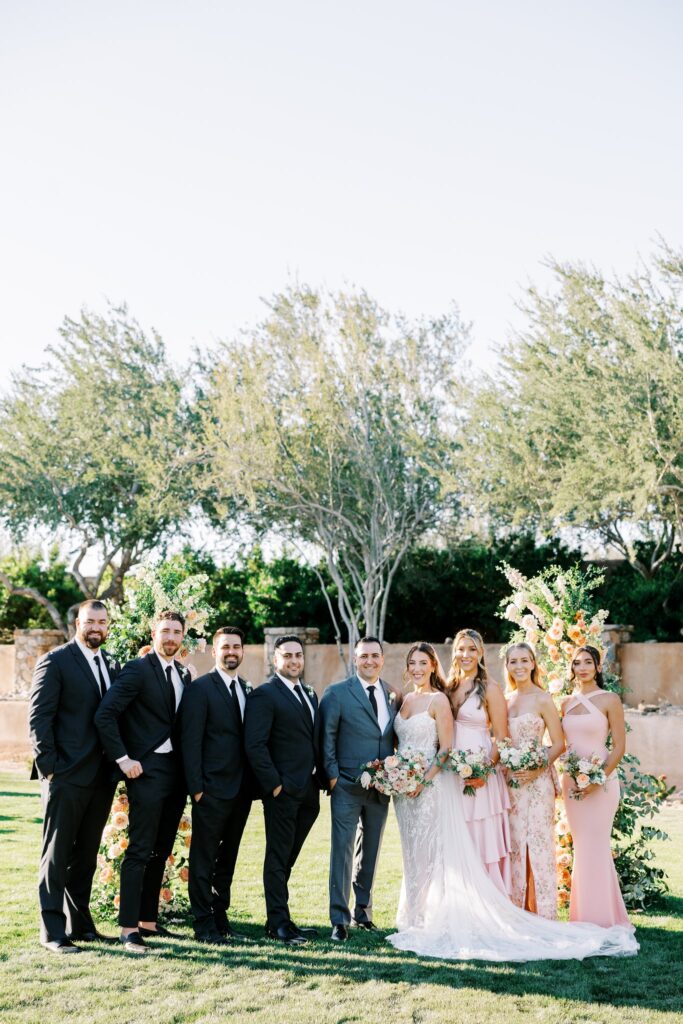 Bridal party standing in row with groomsmen in black suits and groom in gray suit and bridesmaids in light pink dresses holding bouquets.