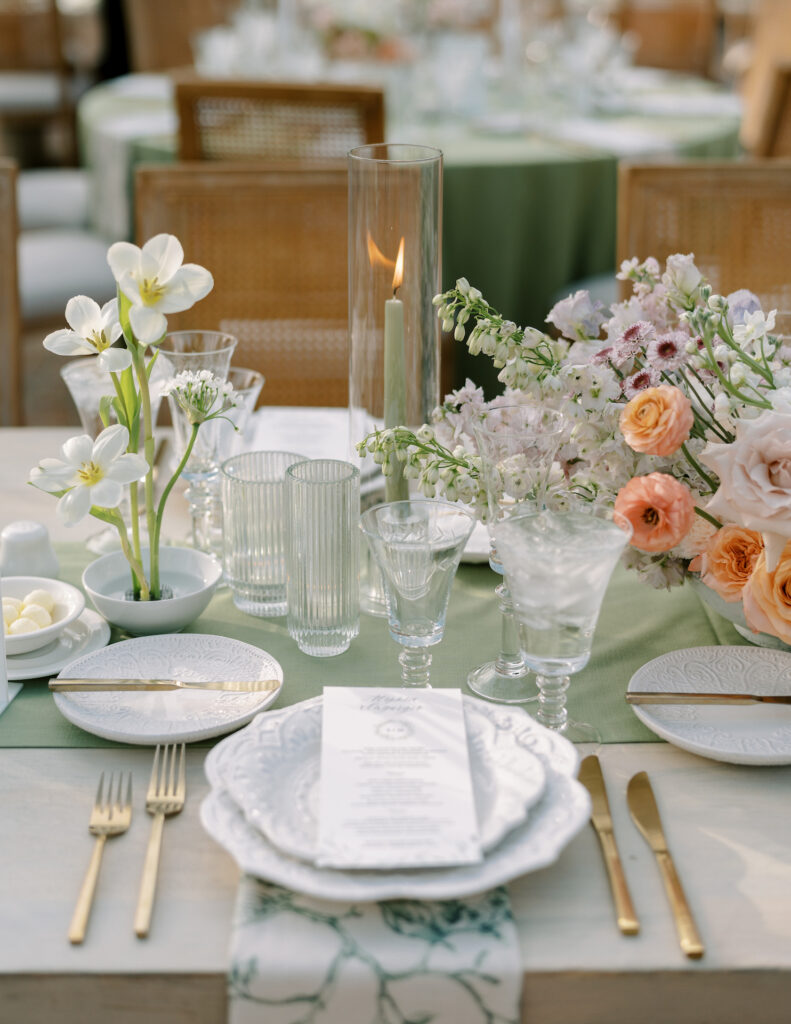 Table scape of wedding reception table with sage green table cloth, floral centerpiece, and white taper candles and white plates.