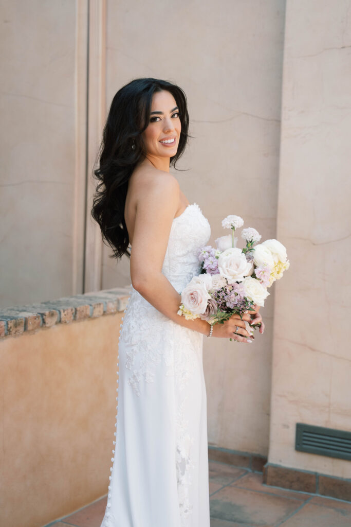 Bride smiling and looking over shoulder holding bouquet of white and purple flowers.