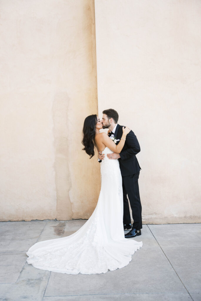 Bride and groom kissing and embrassing in front of tall stucco wall.