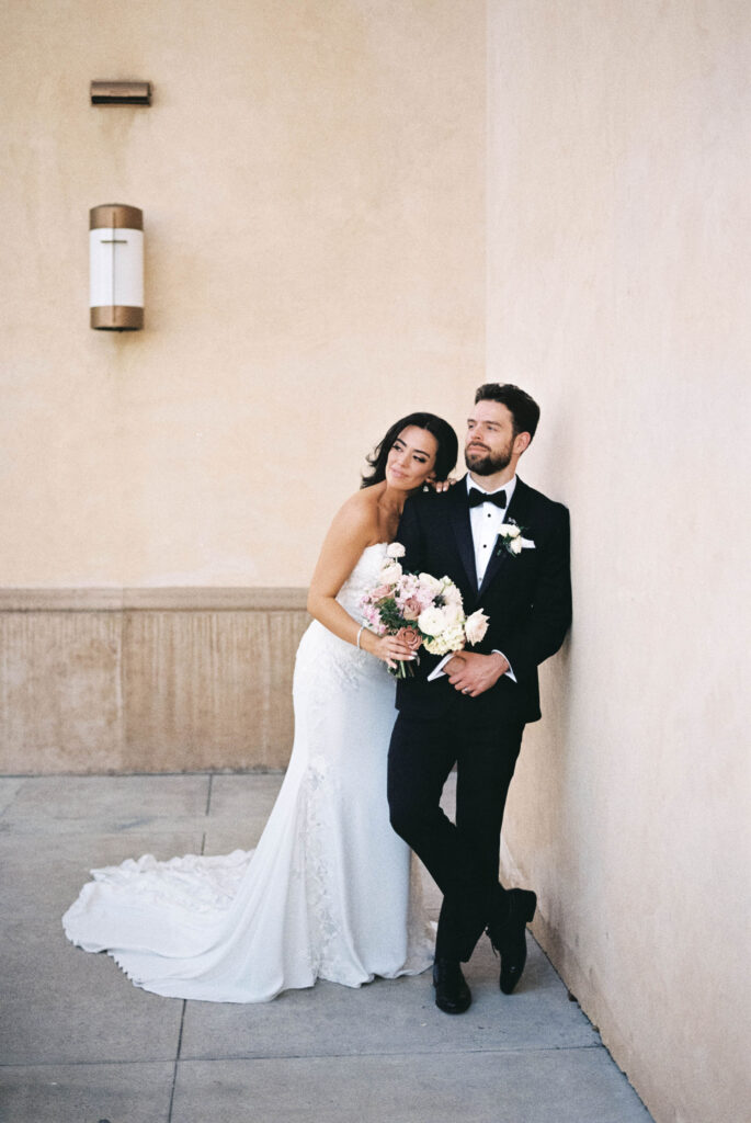 Groom leaning against exterior wall with bride leaning against him holding a bouquet, both looking off to side.