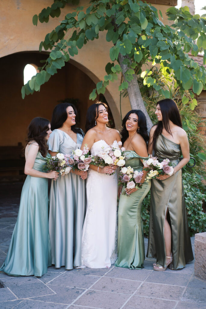 Bride with bridesmaids in shades of green dresses standing in a line, all holding bouquets.