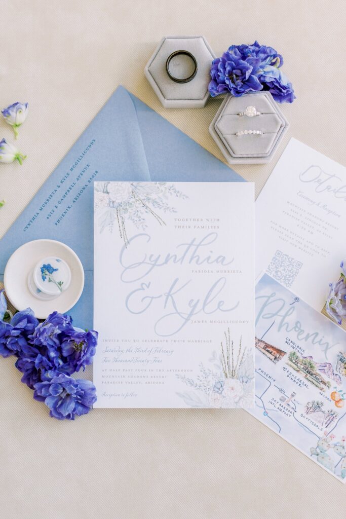 Invitation flat lay of white and blue color palette with wedding rings and floral details.