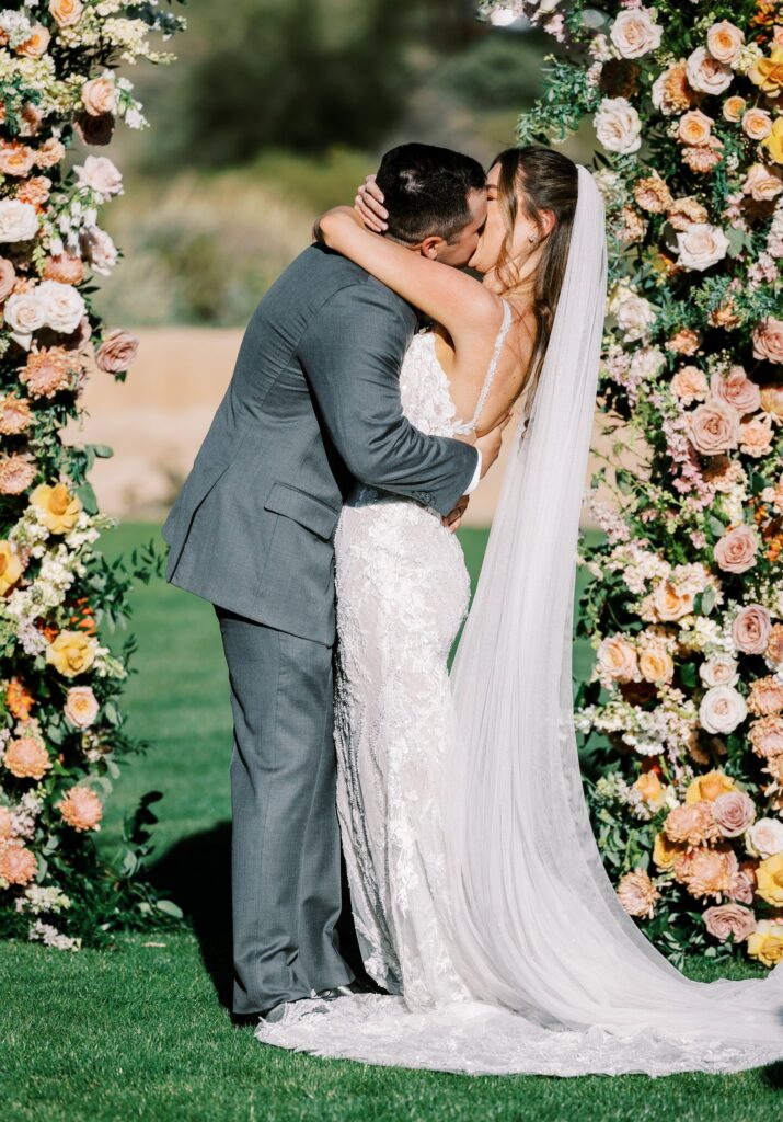 Bride and groom kissing at wedding ceremony between floral pillars.