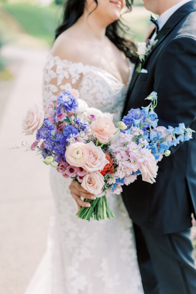 Bride and groom embracing, bride holding out bouquet to side of white, pink, lavender, and blue flowers.