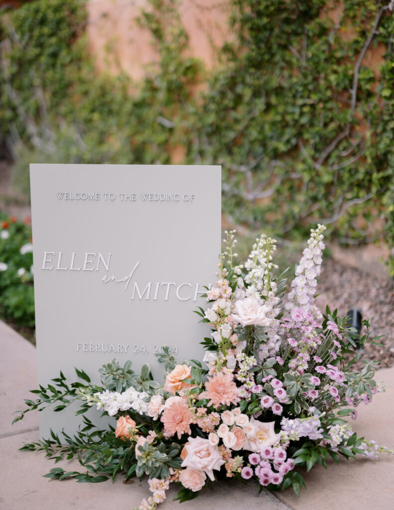 Floral arrangement on ground next to gray wedding welcome sign of purple, peach, pink, and white flowers.