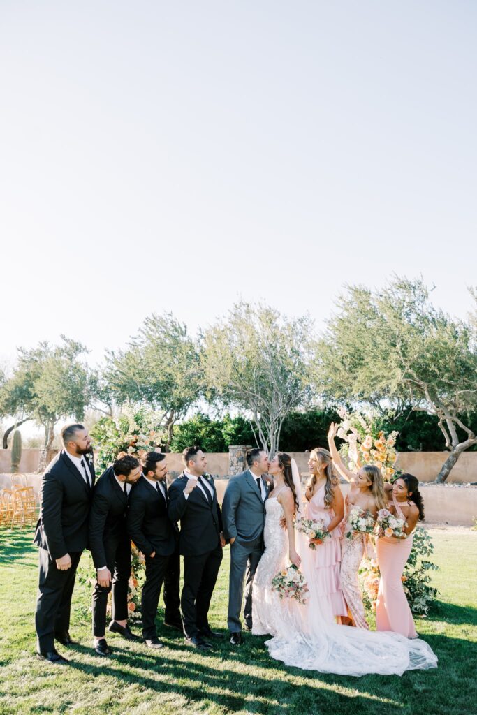 Wedding party celebrating around bride and groom kissing.