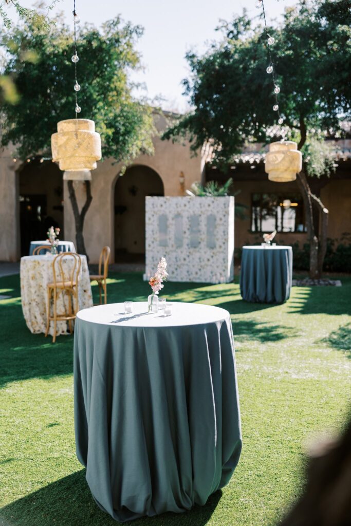 Cocktail hour high boy table with bud vases of flowers with escort board in distance in front of building.