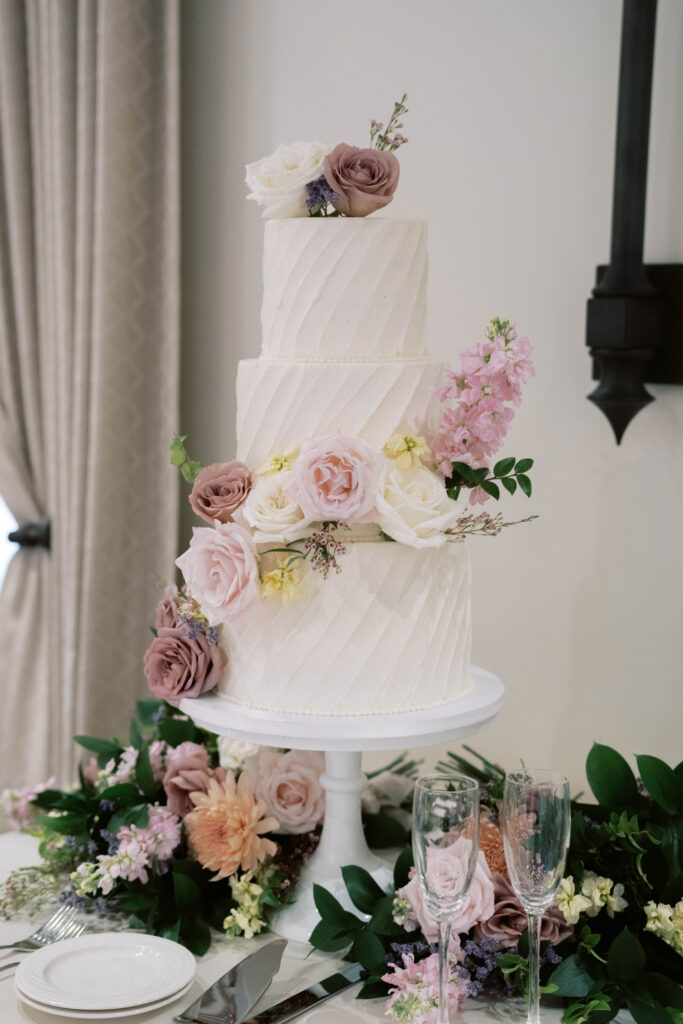 Three tiered white textured wedding cake with added pink, white, yellow, and mauve flowers.