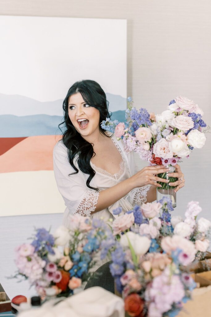 Bride looking to side, with look of surprise on her face, while holding up bouquet of flowers in glass vase.