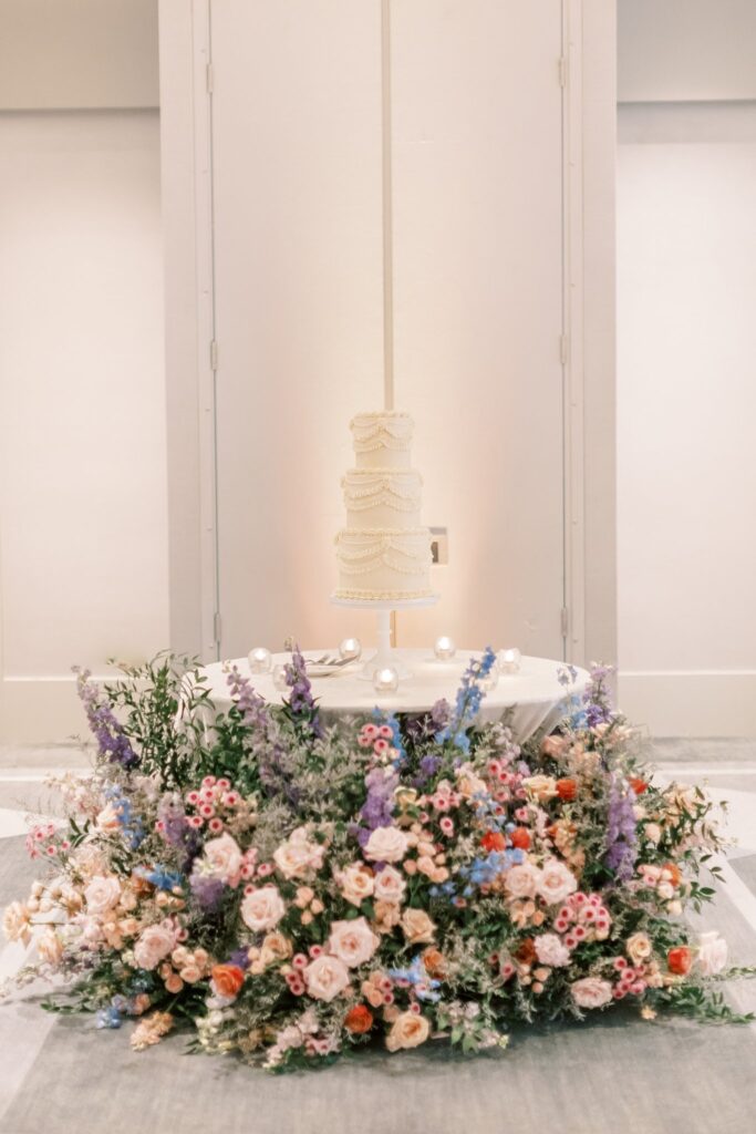 Cake table at inside reception with three tiered white wedding cake on stand resting on table and large ground floral around table.