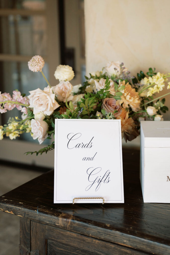 Card and gift table sign at wedding with floral arrangment behind it on table.