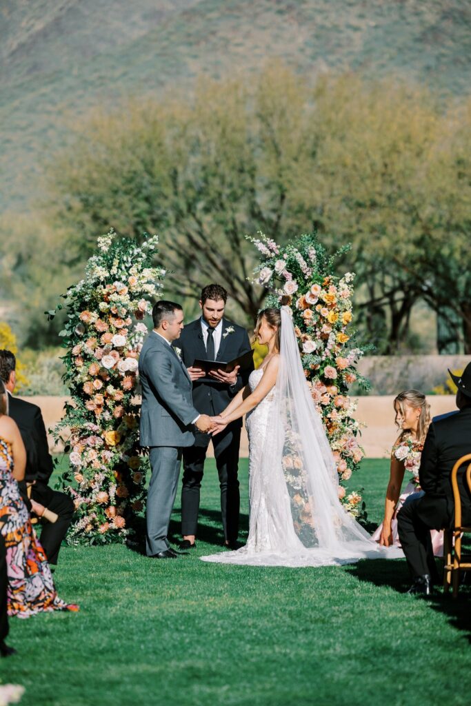 Bride and groom holding hands during wedding ceremony in altar space with officiant behind them and two floral pillars on either side of them.