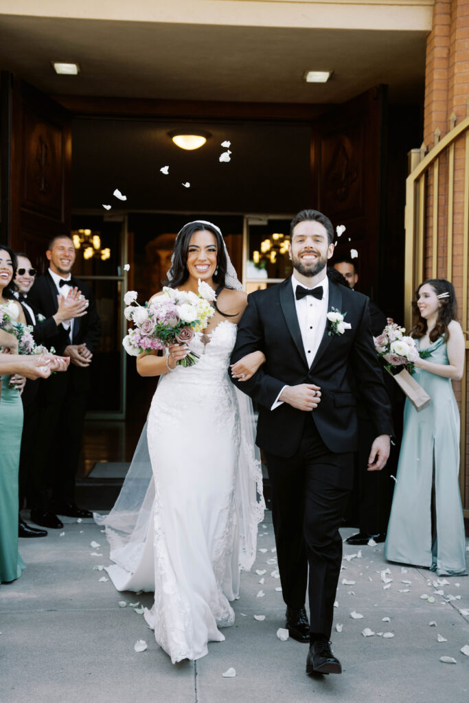 Bride and groom exiting building, smiling, with people lined up on both sides of walkway clapping.