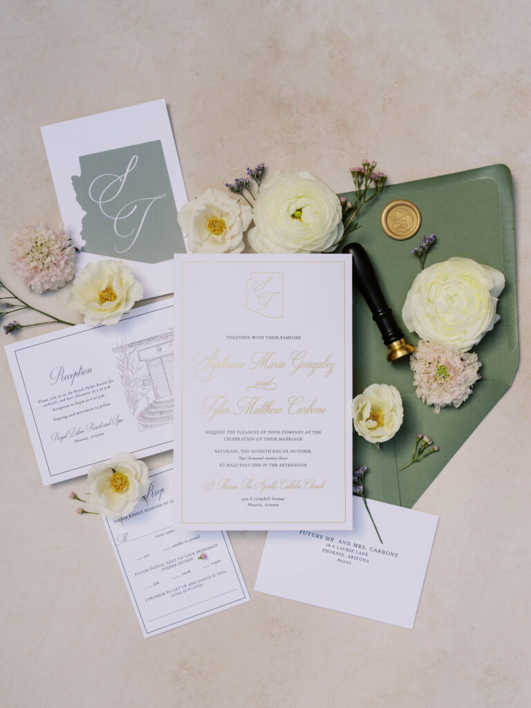 Wedding invitation flat lay in white and sage green colors with flowers scattered around them.