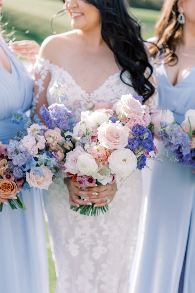 Bouquet of white, pink, lavender, and blue flowers held by bride and bridesmaids in soft blue dresses.