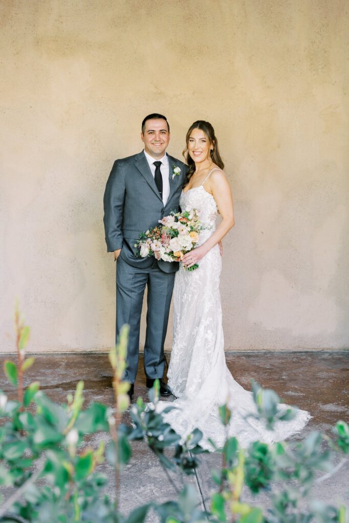 Bride and groom standing close, smiling, in front of stucco wall. Bride holding bouquet.