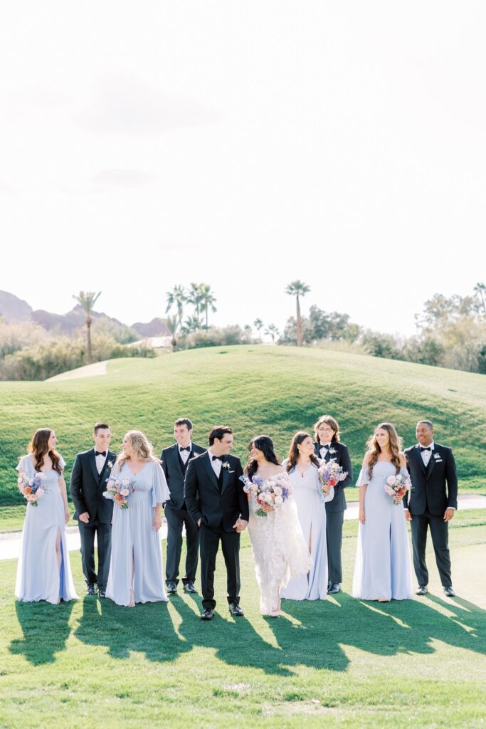 Wedding party walking through grass field with bride and groom in center, all smiling.