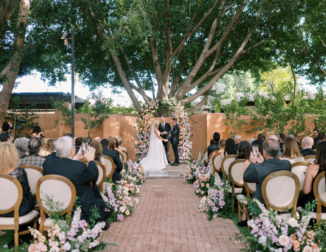 Outdoor ceremony space at Royal Palms with floral arrangements along aisle ground and floral arch in altar space, with bride and groom standing under it with officiant.