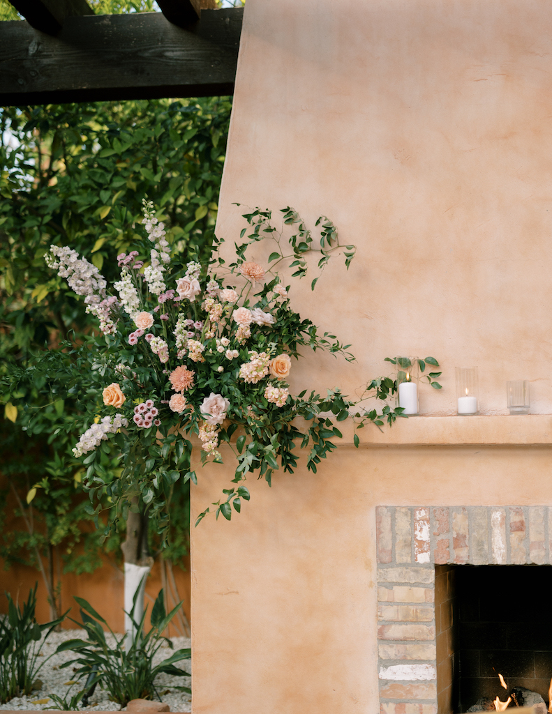 Floral arrangement on fireplace corner of greenery with white, peach, and purple flowers. Pillar candles also on mantle.