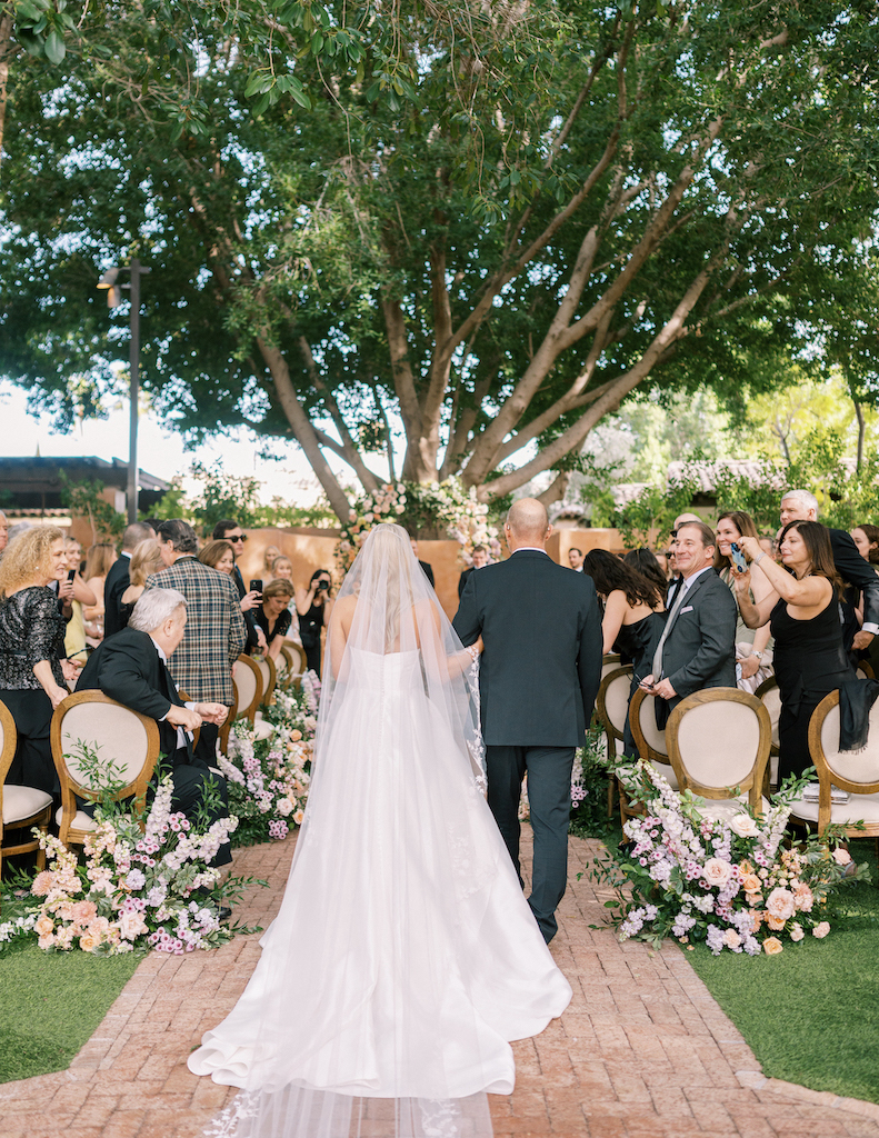 Bride walking down aisle arm in arm with man at outdoor wedding ceremony.