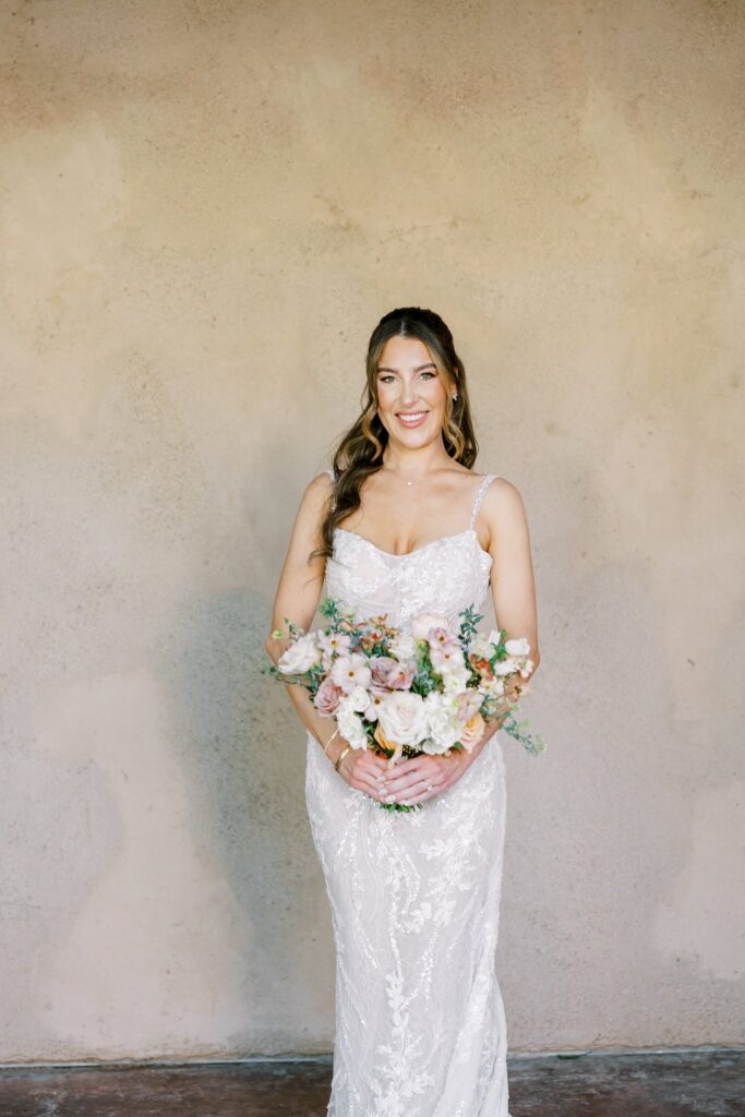 Bride standing, smiling, in front of stucco wall holding bouquet of white and pink flowers.