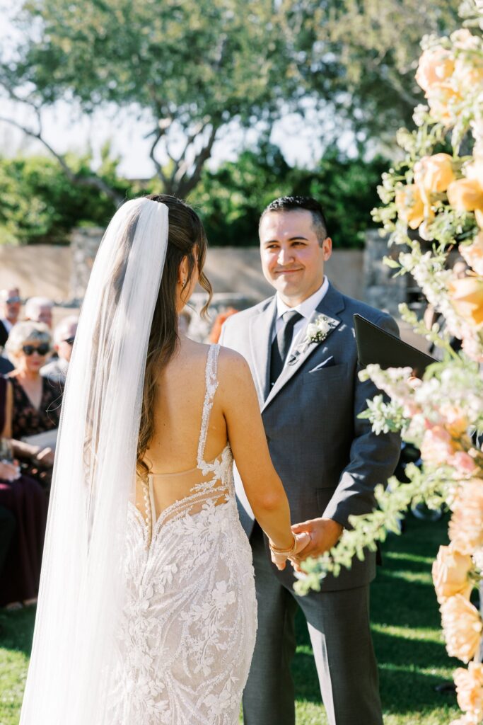 Groom smiling at bride and holding each other's hands standing in altar space at ceremony.