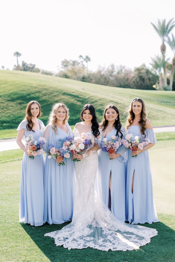 Bouquet of white, pink, lavender, and blue flowers held by bride and bridesmaids in soft blue dresses.