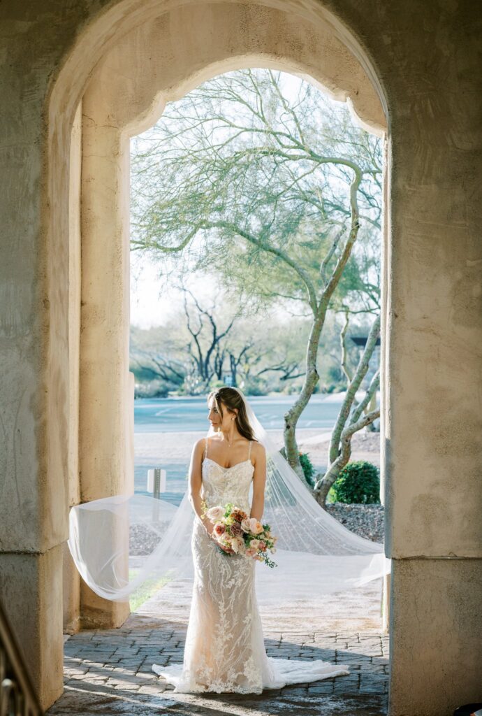 Bride standing in large archway with veil flowering behind her, looking off to side and holding bridal bouquet.