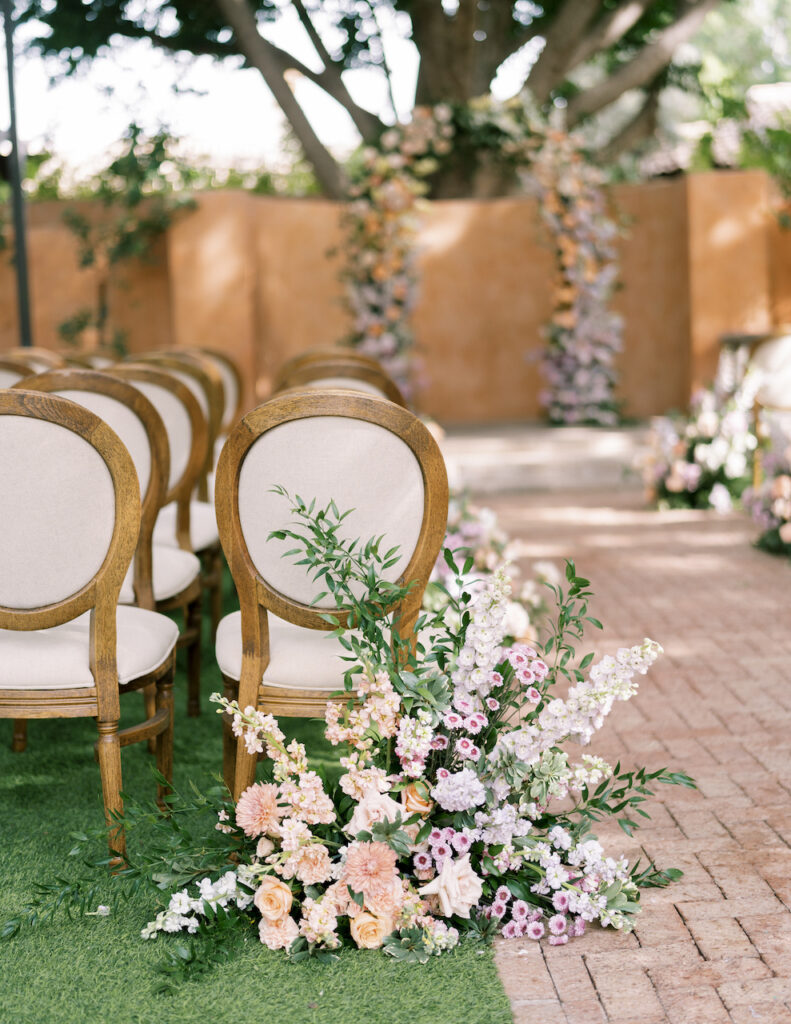 Ground floral arrangement next to aisle chair with purple, pink, and white flowers with greenery.