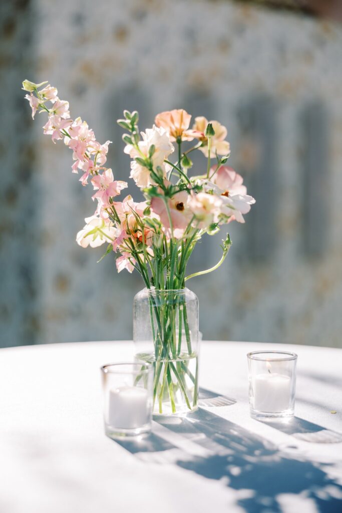 Pink and white flowers in glass bud vase with votive candles next to it.