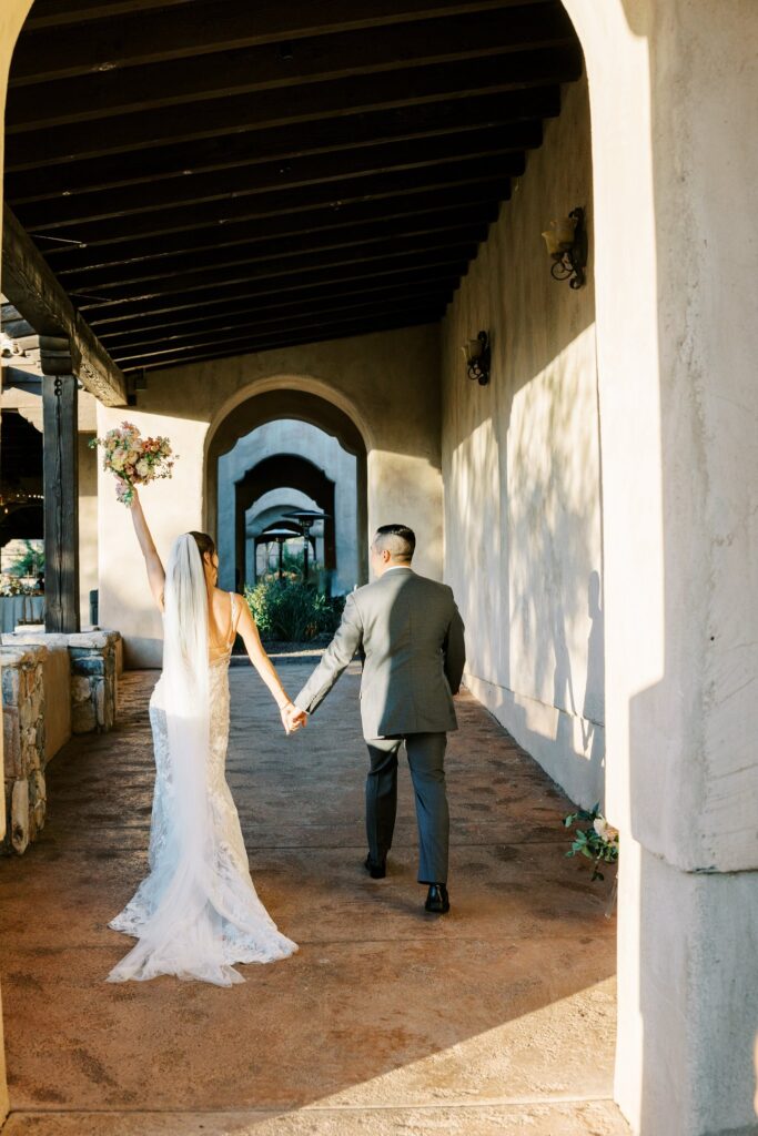 Bride and groom walking away under breezeway holding hands, bride holding up bouquet.