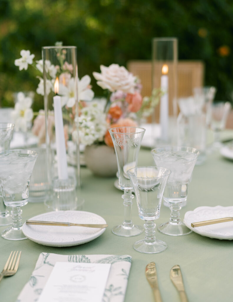 Table scape of wedding reception table with sage green table cloth, floral centerpiece, and white taper candles.