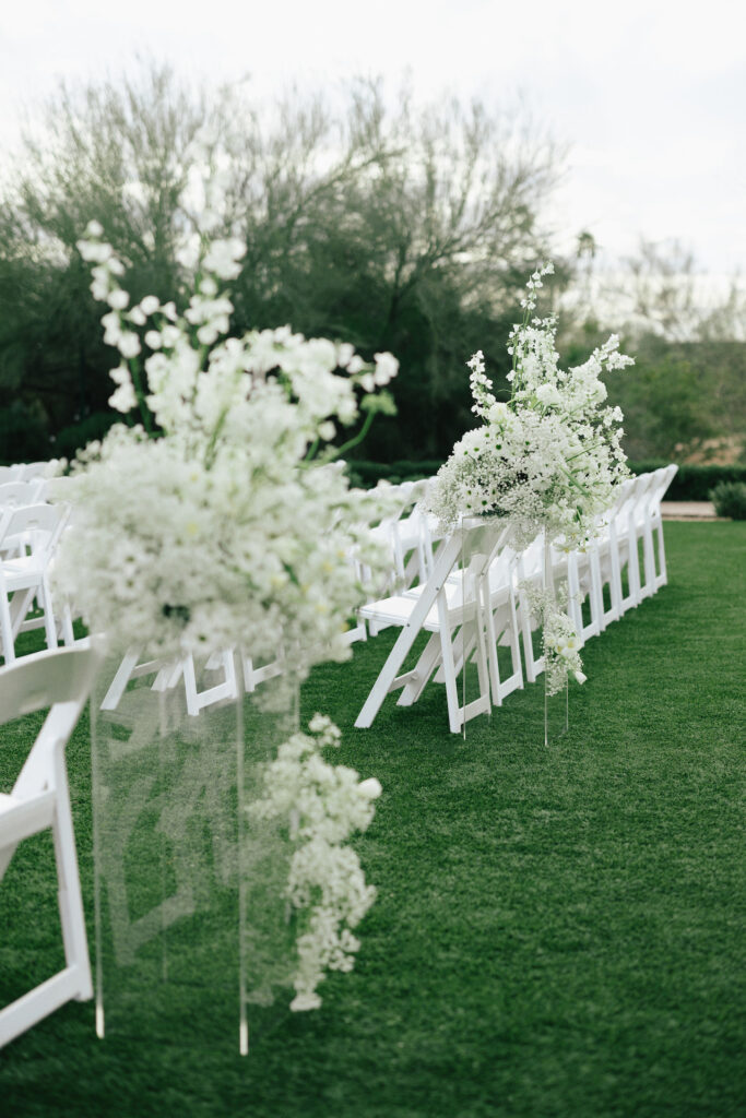 Back of outdoor wedding ceremony aisle with white flower arrangements on clear acrylic pillars and on ground on either side.