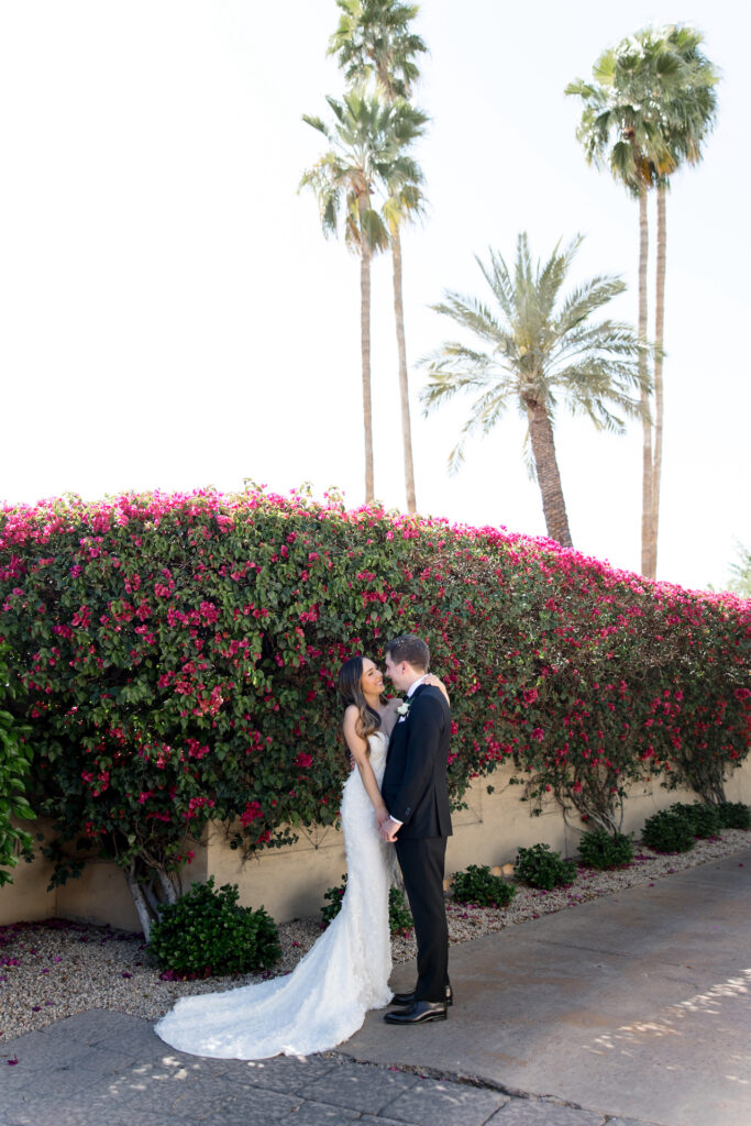 Bride and groom standing close together next to wall covered with bougainvillea with palm trees in the distance.