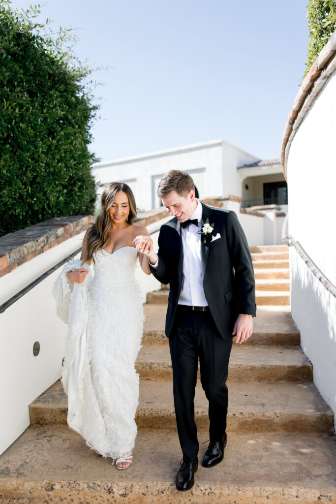 Groom holding bride's hand walking down steps, bride holding up train of dress over arm.