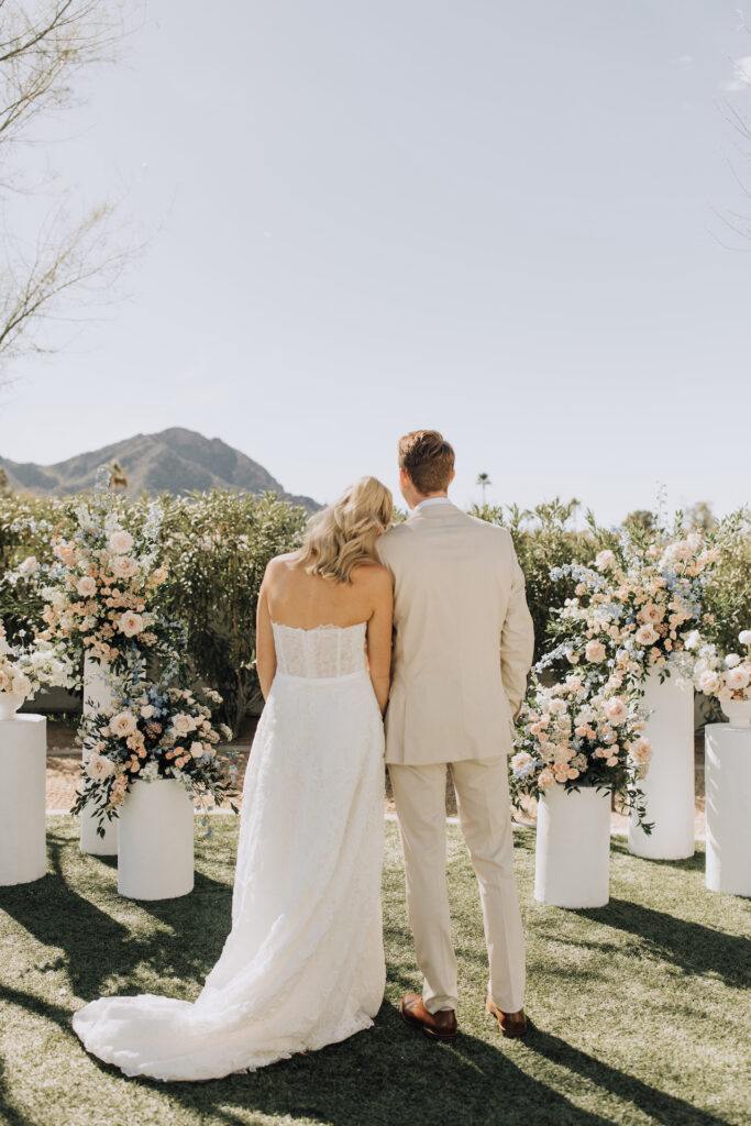 The back of bride and groom in wedding ceremony altar space looking at Camelback Mountain with bride's head resting on groom's shoulder.