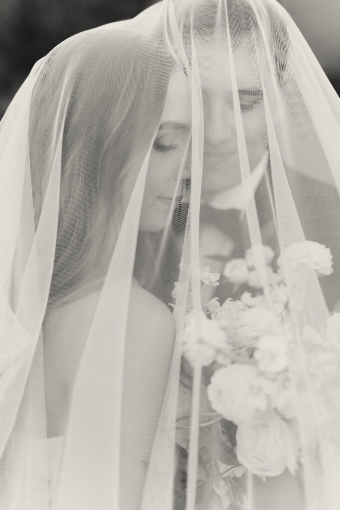 Bride and groom under her veil, gently smiling and looking down at bridal bouquet.