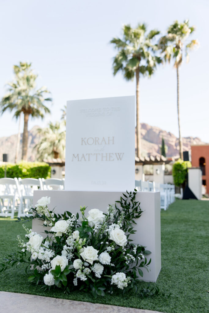 Welcome sign at wedding ceremony with bride and groom's name and ground floral arrangement in front of it.