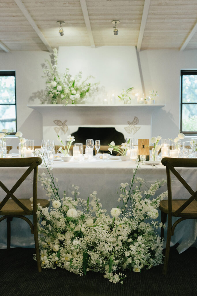 Indoor reception bride and groom table with ground floral and candles and floral on indoor fireplace mantle.