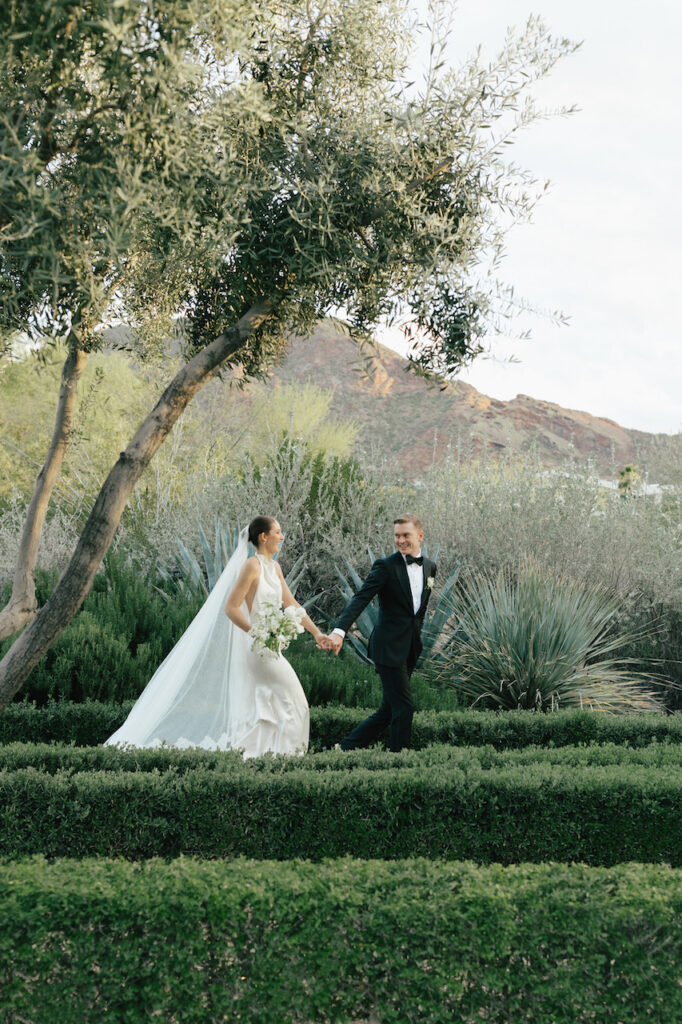 Bride and groom walking in desert landscape holding hands with Camelback Mountain in backdrop.