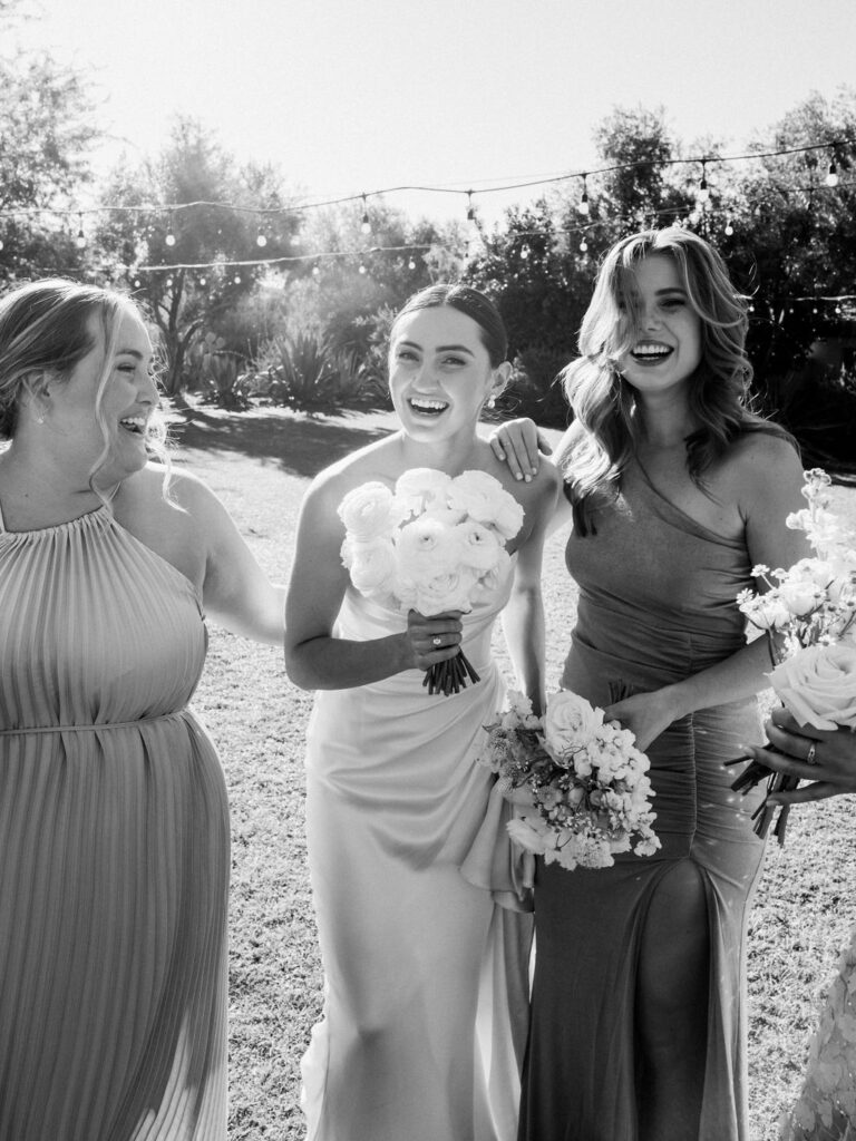 Bride laughing with bridesmaids outside, all holding bouquets.