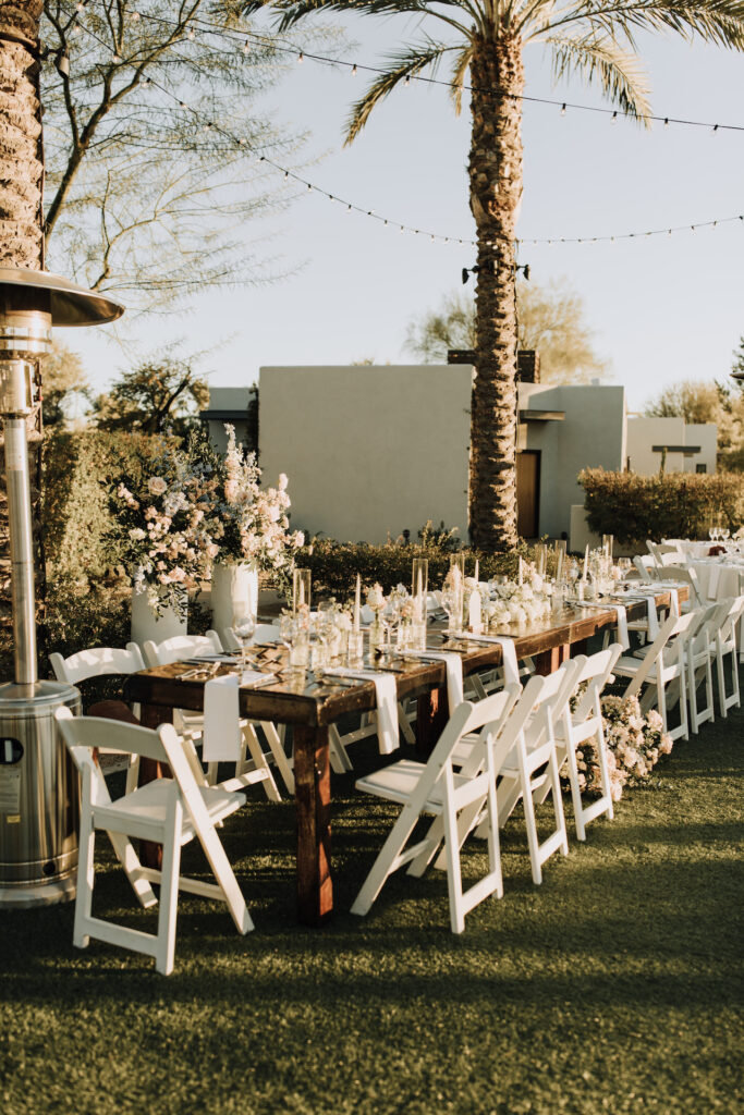 Long outdoor reception table with white chairs and taper candles with bud vases down center.