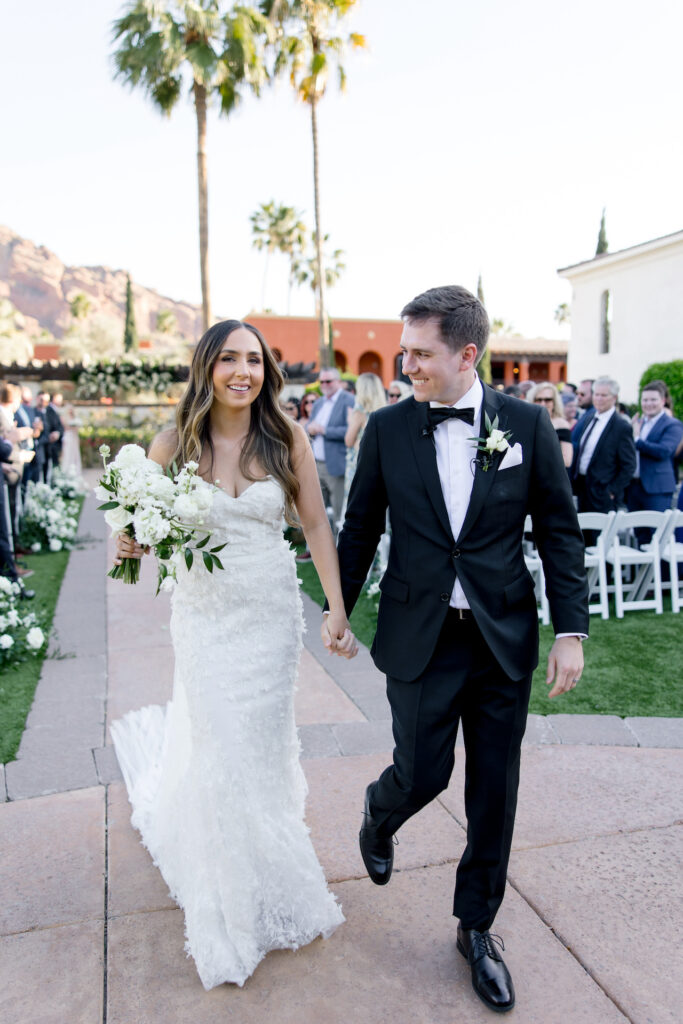 Bride and groom holding hands, exiting wedding ceremony aisle, smiling.