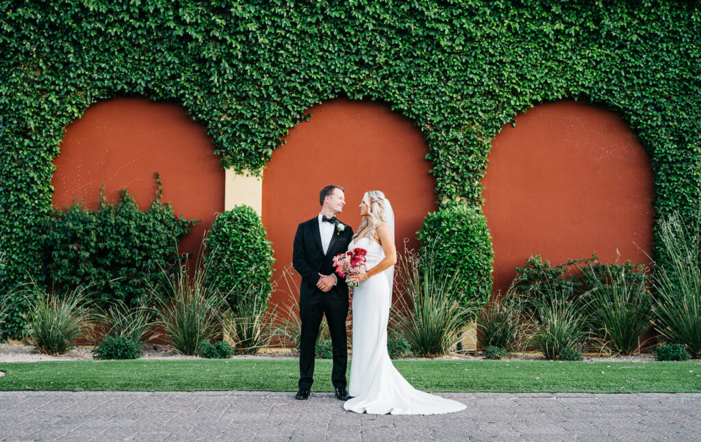 Bride and groom standing next to each other, smiling, bride holding bouquet standing in front of wall with lush greenery growing up wall.